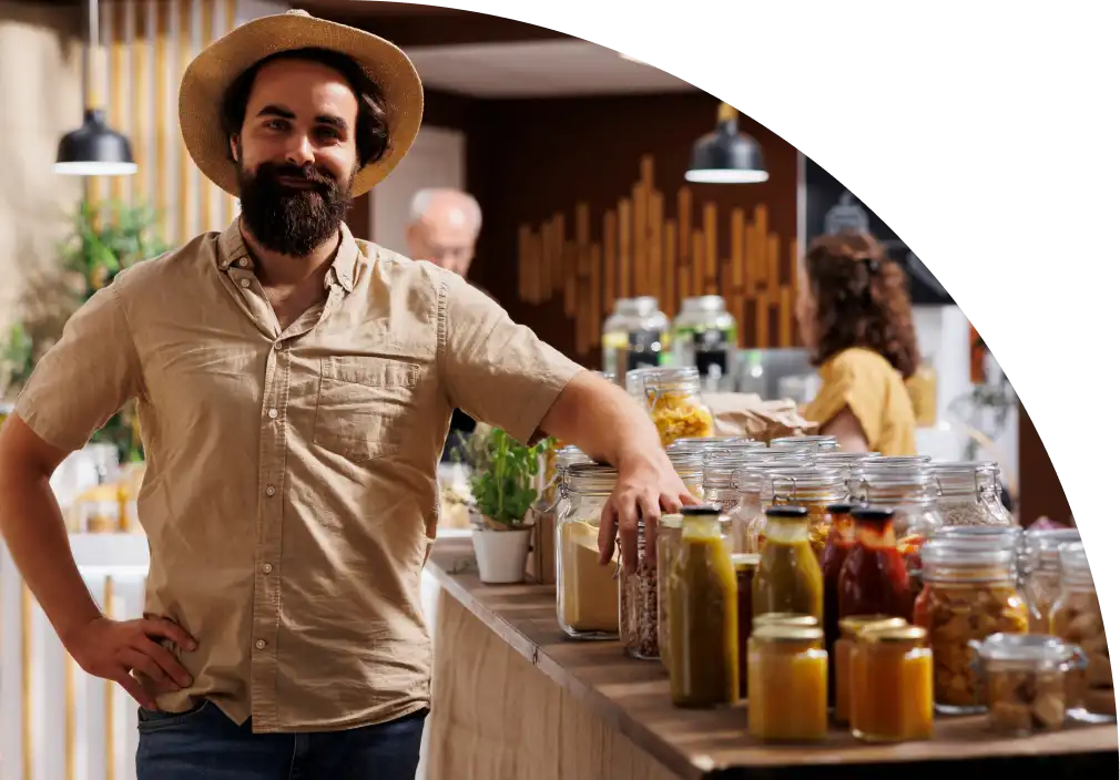 Een man met een baard en een strohoed glimlacht in een rustieke winkel met potten zelfgemaakte jam. Warme verlichting en groen zorgen voor een gastvrije sfeer.