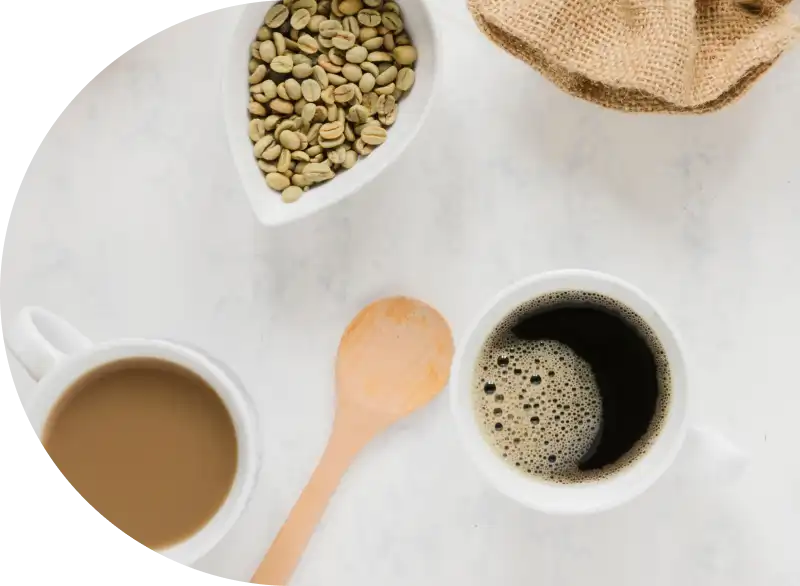 Luchtfoto van een koffieopstelling op een wit oppervlak. Bevat zwarte koffie, koffie met melk, gemalen koffie, groene bonen, een jutezak en een houten lepel.